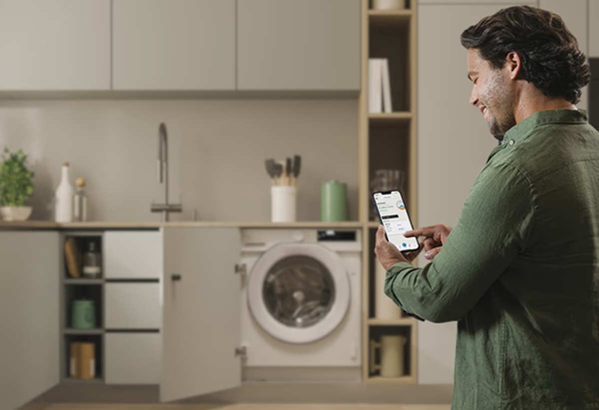 Απεικονίζεται άνδρας να χρησιμοποιεί την εφαρμογή hOn. Φαίνεται το πλυντήριο στο βάθος.